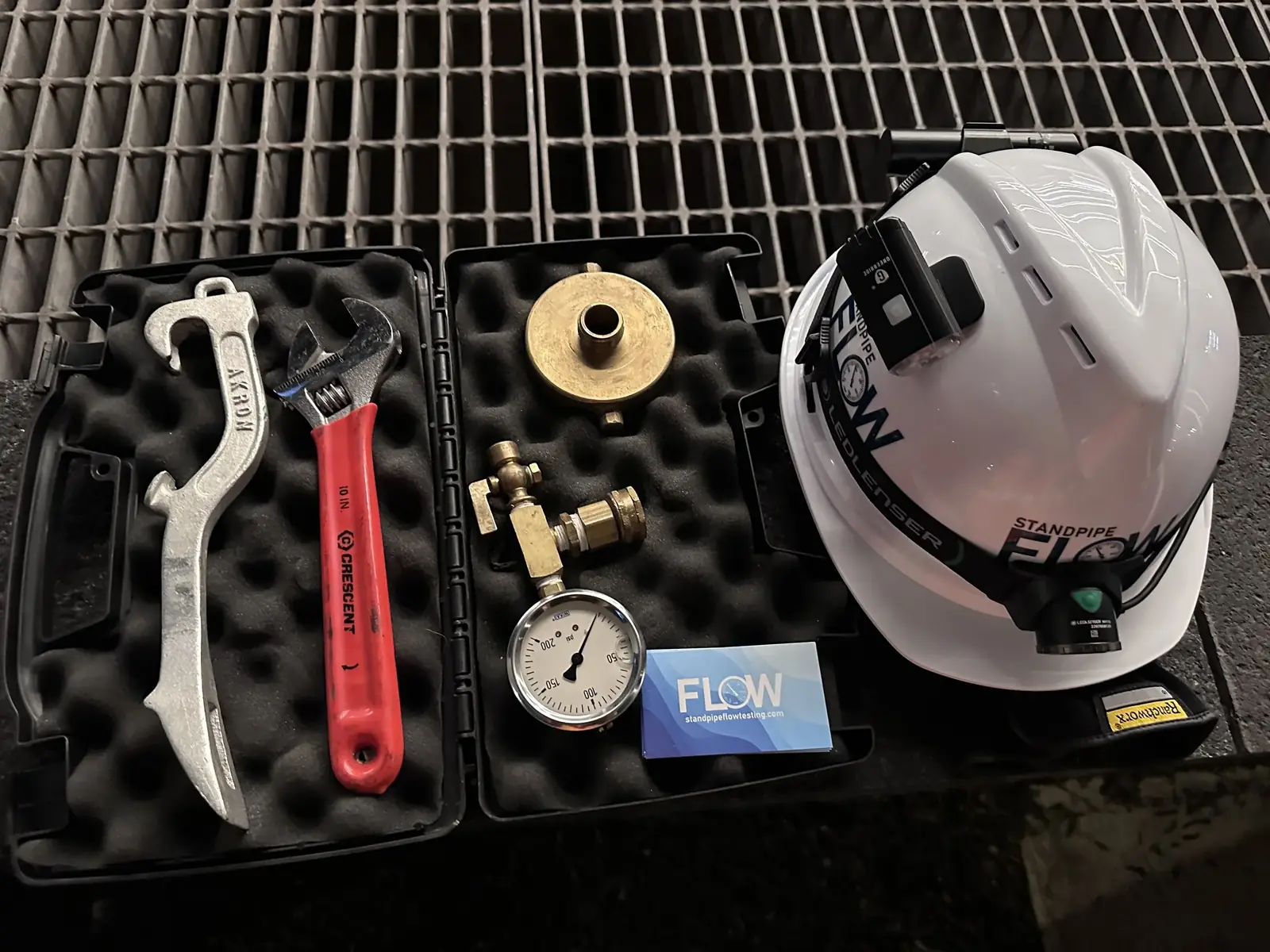 Open black tool case on metal grating holding a hydrant wrench, a red adjustable wrench, a brass fitting, a pressure gauge and a FLOW business card, next to a white FLOW hard hat with a headlamp