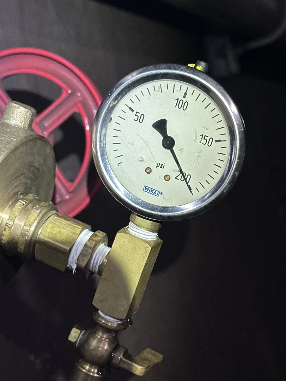 Close-up of a round pressure gauge with a white dial and black needle, mounted on a brass fitting beside a red valve handle