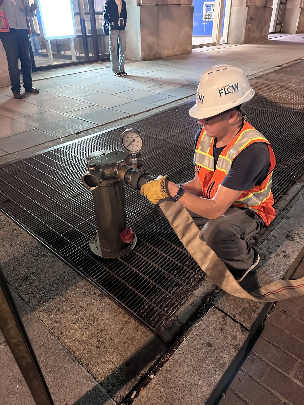FLOW technician in a hi-vis vest and gloves at a sidewalk standpipe connection fitted with a test pressure gauge