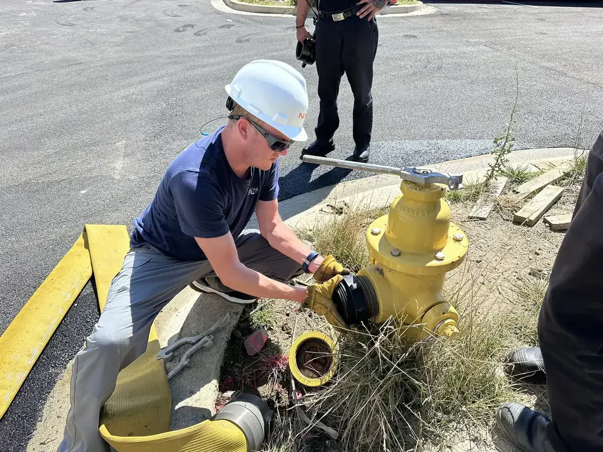 FLOW technician in a white hard hat connecting a fire hose to the outlet of a yellow fire hydrant at a curbside flow test location